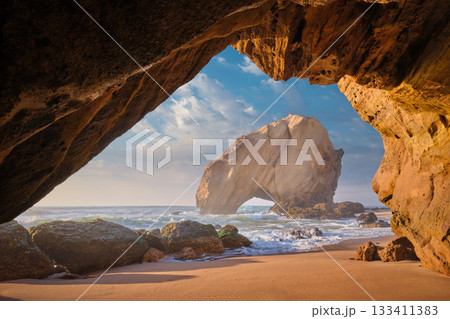 Penedo do Guincho, a large boulder rock arch at Praia da Santa Cruz, Portugal, with ocean waves and sandy beach as seen from grotto cave on sunset 133411383