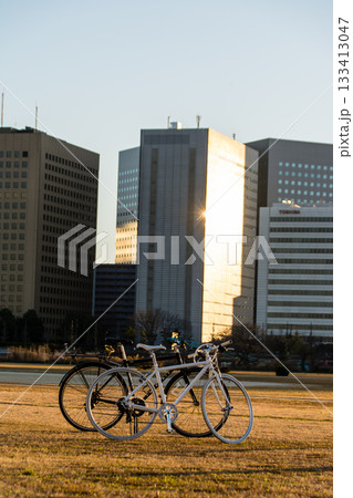 多摩川の河川敷に置かれた白と黒の自転車と川向うの川崎に見える夕日を浴びてオレンジ色になったビル群 133413047