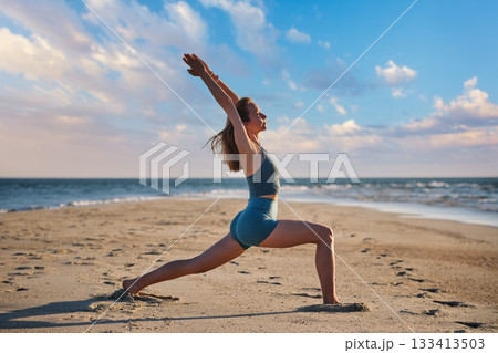 A woman performs Virabhadrasana 1, or Warrior 1 pose, on the beach during sunset. She stands strong against the backdrop of the ocean and colorful sky, embracing tranquility and strength. A woman performs Virabhadrasana 1, or Warrior 1 pose, on the beach during sunset. She stands strong against the backdrop of the ocean and colorful sky, embracing tranquility and strength. 133413503