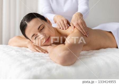 Young female smiling softly while receiving relaxing shoulder massage at spa Young female smiling softly while receiving relaxing shoulder massage at spa 133414955