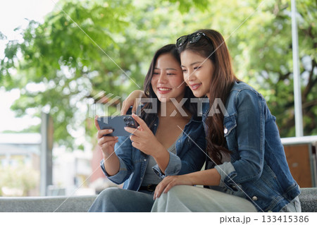 Friendship and Connection. Two young women enjoying a moment together while watching a video on a smartphone outdoors. 133415386