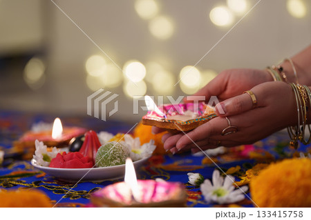 Diwali Ritual. Hand holding a lit diya amidst festive decorations. 133415758