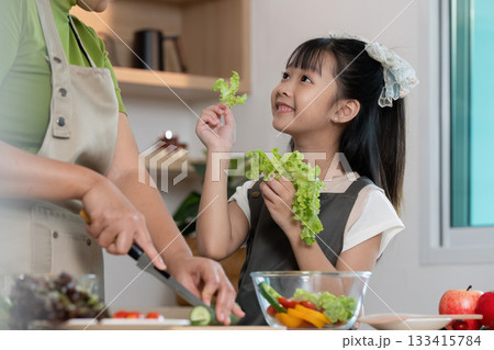 Family Cooking. Child enjoying fresh lettuce while parent prepares salad in modern kitchen. 133415784