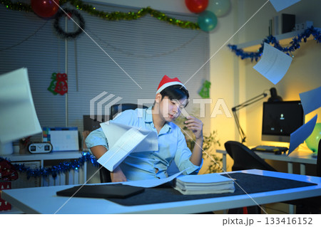 tired man with sad face sitting at desk in office or home office, many stacks of papers and documents on work desk, Christmas tree in background. overtime work and reports during holidays 133416152