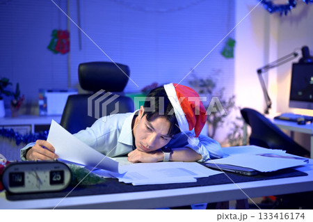 tired man with sad face sitting at desk in office or home office, many stacks of papers and documents on work desk, Christmas tree in background. overtime work and reports during holidays tired man with sad face sitting at desk in office or home office, many stacks of papers and documents on work desk, Christmas tree in background. overtime work and reports during holidays 133416174
