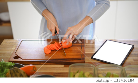 Closeup of young female hands chopping fresh orange carrot on board while in modern kitchen Closeup of young female hands chopping fresh orange carrot on board while in modern kitchen 133416194