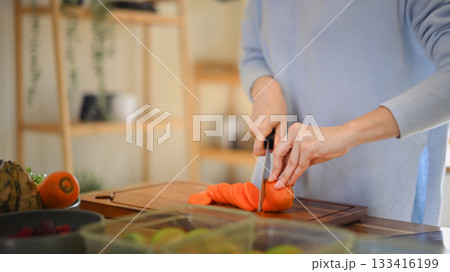 Closeup of young female hands chopping fresh orange carrot on board while in modern kitchen Closeup of young female hands chopping fresh orange carrot on board while in modern kitchen 133416199