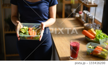 A woman holding a food container with fresh vegetables and a smoothie on the table, symbolizing a healthy and mindful lifestyle at home A woman holding a food container with fresh vegetables and a smoothie on the table, symbolizing a healthy and mindful lifestyle at home 133416200