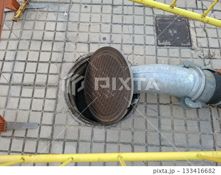 Water is being pumped out of a manhole in a busy city street. Surrounding the area are safety barriers, highlighting the ongoing maintenance and construction effort. Water is being pumped out of a manhole in a busy city street. Surrounding the area are safety barriers, highlighting the ongoing maintenance and construction effort. 133416682