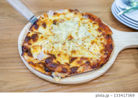 A person uses a knife to cut a slice of thin crust pizza on a round wooden board, showing melted cheese and pineapple pieces on top. A person uses a knife to cut a slice of thin crust pizza on a round wooden board, showing melted cheese and pineapple pieces on top. 133417369