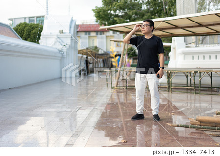 A man pauses to adjust his glasses while standing in a temple courtyard, blending modern casual style with a mindful, reflective presence. A man pauses to adjust his glasses while standing in a temple courtyard, blending modern casual style with a mindful, reflective presence. 133417413