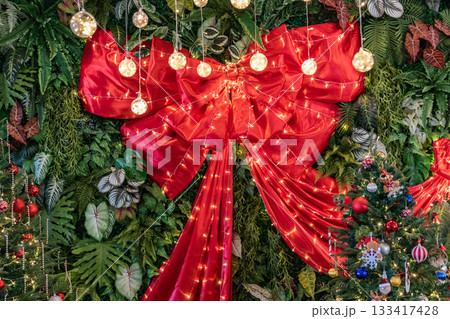 A large red ribbon with multiple folds rests against a wall of green leaves, with warm string lights hanging above and running across the fabric. 133417428