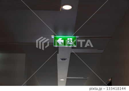 A green fire escape sign hangs from the ceiling, showing an exit sign green light box with a running man and an arrow pointing right for safety and evacuation. A green fire escape sign hangs from the ceiling, showing an exit sign green light box with a running man and an arrow pointing right for safety and evacuation. 133418144