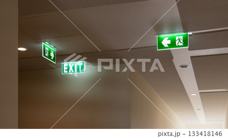 A green fire escape sign hangs from the ceiling, showing an exit sign green light box with a running man and an arrow pointing right for safety and evacuation. 133418146