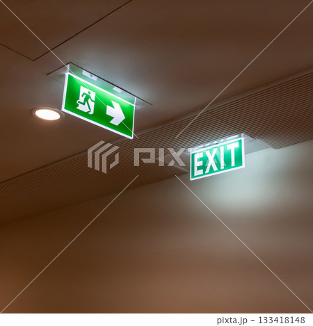 A green fire escape sign hangs from the ceiling, showing an exit sign green light box with a running man and an arrow pointing right for safety and evacuation. 133418148