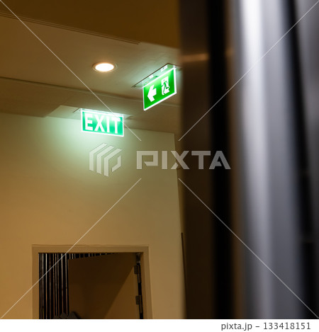A green fire escape sign hangs from the ceiling, showing an exit sign green light box with a running man and an arrow pointing right for safety and evacuation. 133418151