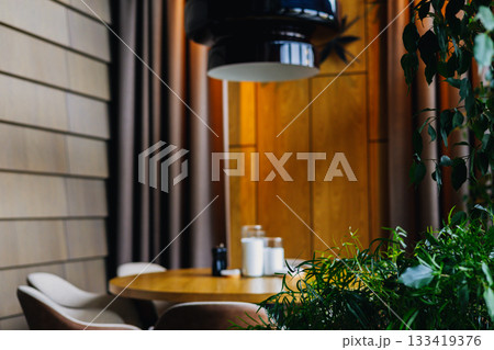 An inviting restaurant interior with a round wooden table, stylish chairs decorative elements, complemented by lush green plants and a modern lighting An inviting restaurant interior with a round wooden table, stylish chairs decorative elements, complemented by lush green plants and a modern lighting 133419376