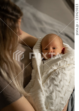 A mother lovingly holds her baby wrapped snugly in a soft blanket, both enjoying a peaceful moment in a calm and warm setting. A mother lovingly holds her baby wrapped snugly in a soft blanket, both enjoying a peaceful moment in a calm and warm setting. 133423013