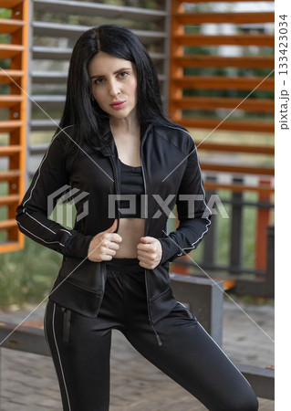 athletic brunette girl stands confidently in fitted workout gear at a park. The scene captures her determination and strength in a vibrant outdoor setting during the day. 133423034
