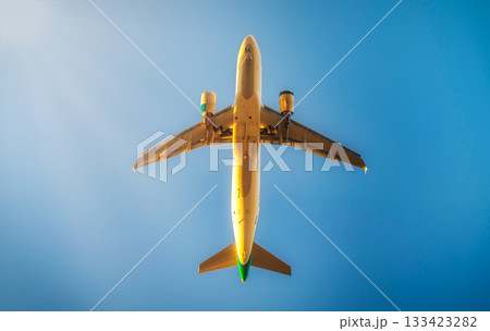 The image shows an airplane captured from below, with its underbelly and landing gear clearly visible against a clear blue sky. The sunlight is casting a glow on parts of the plane, creating a few The image shows an airplane captured from below, with its underbelly and landing gear clearly visible against a clear blue sky. The sunlight is casting a glow on parts of the plane, creating a few 133423282