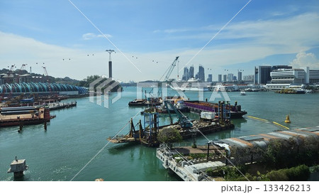 A bustling waterfront construction site with cranes, cable cars, vessels, and a distant cruise ship showcases an active industrial zone blending maritime activity with a modern urban skyline. 133426213