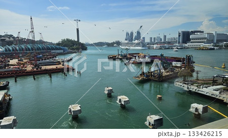 A bustling waterfront construction site with cranes, cable cars, vessels, and a distant cruise ship showcases an active industrial zone blending maritime activity with a modern urban skyline. A bustling waterfront construction site with cranes, cable cars, vessels, and a distant cruise ship showcases an active industrial zone blending maritime activity with a modern urban skyline. 133426215