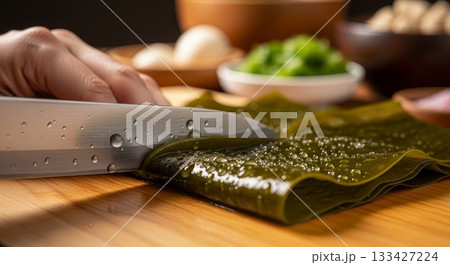 Professional chef hands slicing fresh green kelp seaweed with sharp knife on wooden cutting board in modern kitchen with water droplets 133427224