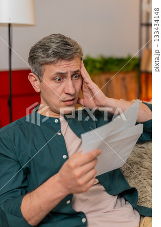 Young man reacts with stress and worry after opening envelope with credit bill at home indoors 133434148