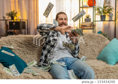Young man throwing dollar bills cash at home spending money playful celebration sudden wealth moment 133434200