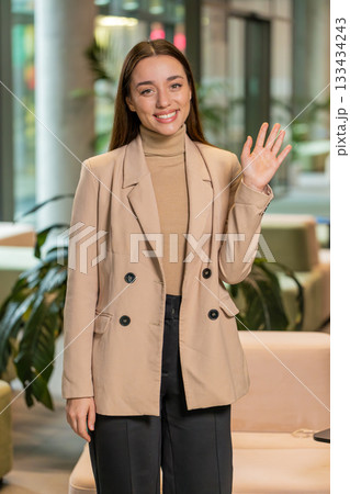 Happy young Caucasian businesswoman looking at camera and waves hand hello in office hotel lobby Happy young Caucasian businesswoman looking at camera and waves hand hello in office hotel lobby 133434243