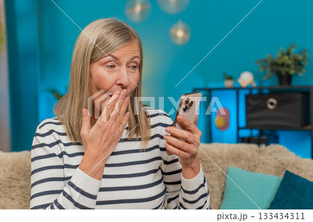 Mature woman celebrating online lottery win victory on smartphone, happily viewing good news at home Mature woman celebrating online lottery win victory on smartphone, happily viewing good news at home 133434311