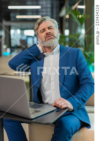 Tired middle-aged businessman suffers from spine neck muscle pain working on laptop in office lobby 133434319