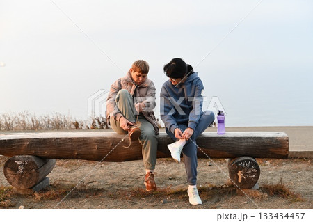 A 50-year-old Russian woman and her 60-year-old friend are sitting on a bench putting on sneakers and tying their laces, getting ready for sports. 133434457