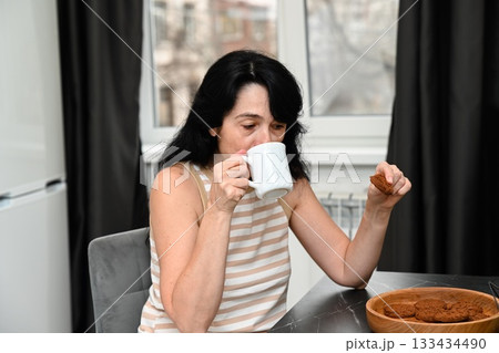 A 50 years old Russian woman takes a sip of coffee with a bitten cookie in hand, enjoying a sleepy gentle morning in a quiet reflective calm mood. 133434490