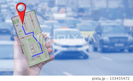 A man's hand holds a smartphone with an online map, on which a destination is marked. In the background, a busy road is shown. The concept of internet navigation and modern technology is represented. 133435472