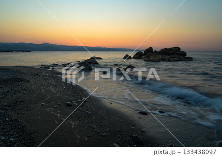 Sunset casts golden hues over tranquil beach with gentle waves lapping at rocky shoreline 133438097