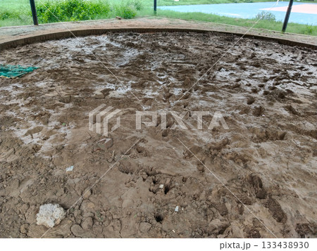 Muddy circular sports arena with wet soil surface and brick boundary in open park, traditional outdoor ground prepared for local games or wrestling, natural earth field surrounded by green landscape 133438930