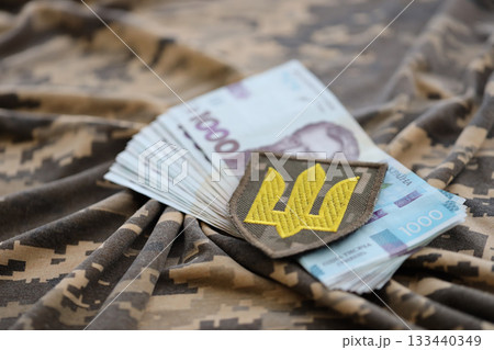 Ukrainian army symbol and bunch of hryvnia bills on military uniform. Payments to soldiers of the Ukrainian army, salaries to the military. War in Ukraine Ukrainian army symbol and bunch of hryvnia bills on military uniform. Payments to soldiers of the Ukrainian army, salaries to the military. War in Ukraine 133440349