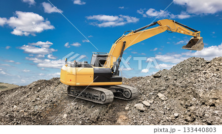 An excavator is moving dirt at a construction site with a clear blue sky and scattered clouds overhead. Piles of gravel and rocks are visible around the machine An excavator is moving dirt at a construction site with a clear blue sky and scattered clouds overhead. Piles of gravel and rocks are visible around the machine 133440803