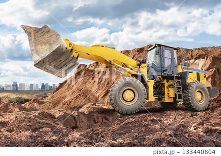 A large yellow construction loader is digging and moving dirt at a building site, with a city skyline visible in the distance and clouds overhead 133440804