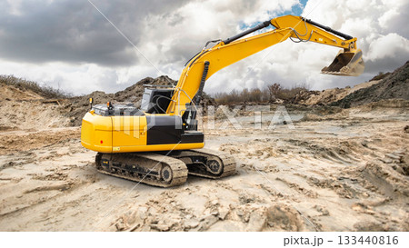 A yellow excavator works on a construction site, digging into the soil under cloudy skies with a landscape of dirt and rocks in the background A yellow excavator works on a construction site, digging into the soil under cloudy skies with a landscape of dirt and rocks in the background 133440816