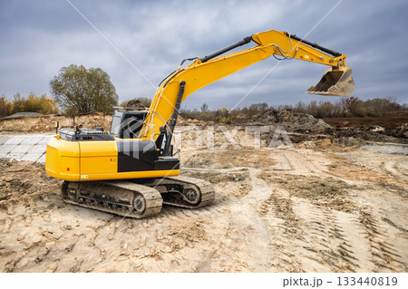 A yellow excavator is working at a construction site in an urban area, moving dirt and preparing the ground under overcast skies A yellow excavator is working at a construction site in an urban area, moving dirt and preparing the ground under overcast skies 133440819