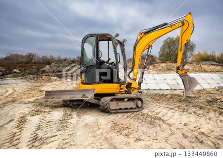 A yellow excavator is positioned on a construction site, preparing to dig into the sandy ground with cloudy skies overhead A yellow excavator is positioned on a construction site, preparing to dig into the sandy ground with cloudy skies overhead 133440860