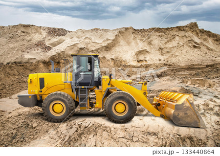 A wheel loader is actively moving dirt at a construction site while surrounded by large mounds of earth under an overcast sky, showcasing heavy machinery in action 133440864