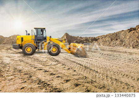 A yellow wheeled bulldozer or loader is working on a construction site, moving dirt and preparing the ground under a clear sky with bright sunlight 133440875