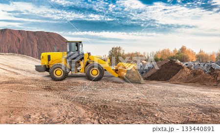 Yellow loader moves earth at a construction site surrounded by hills and trees during sunset, showcasing the machinery's strength and efficiency Yellow loader moves earth at a construction site surrounded by hills and trees during sunset, showcasing the machinery's strength and efficiency 133440891