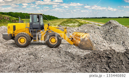 A yellow loader digs into a large gravel mound on a construction site surrounded by green fields under a blue sky, showcasing ongoing earth-moving activities A yellow loader digs into a large gravel mound on a construction site surrounded by green fields under a blue sky, showcasing ongoing earth-moving activities 133440892