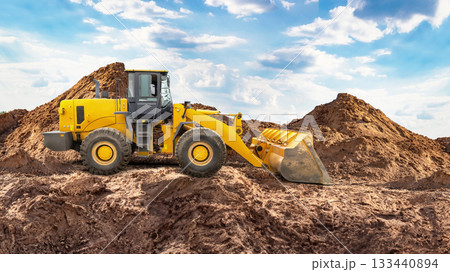 A yellow loader moves dirt at a construction site, with large mounds of earth in the background under a bright blue sky and scattered clouds A yellow loader moves dirt at a construction site, with large mounds of earth in the background under a bright blue sky and scattered clouds 133440894