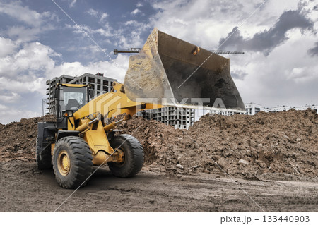 A large bulldozer loader lifts a pile of dirt on a construction site surrounded by buildings and cranes under a cloudy sky during the day A large bulldozer loader lifts a pile of dirt on a construction site surrounded by buildings and cranes under a cloudy sky during the day 133440903