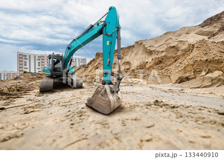 Excavator works on a construction site moving dirt and sand near residential buildings, under a cloudy sky in an urban environment Excavator works on a construction site moving dirt and sand near residential buildings, under a cloudy sky in an urban environment 133440910